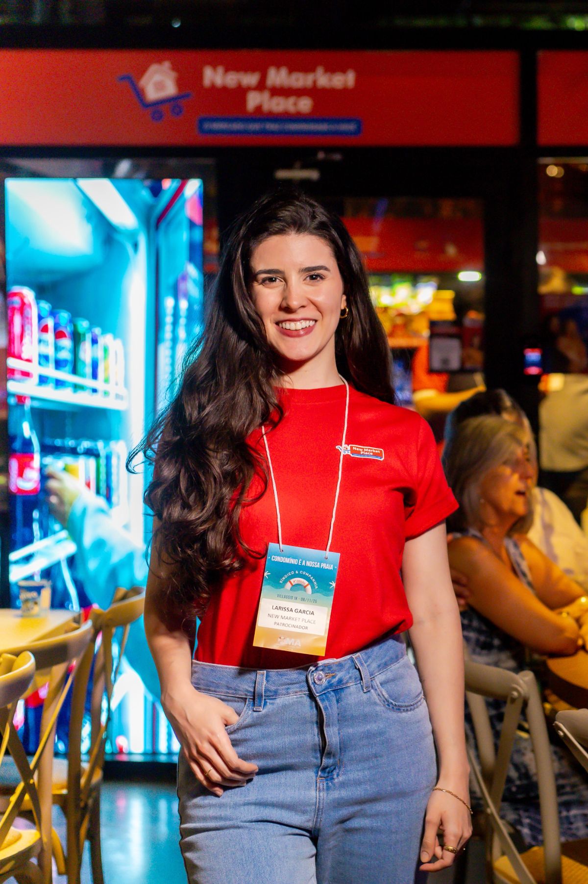 Larissa Garcia, da equipe New Market Place, de camiseta vermelha e crachá, em evento do setor de condomínios, em frente à loja